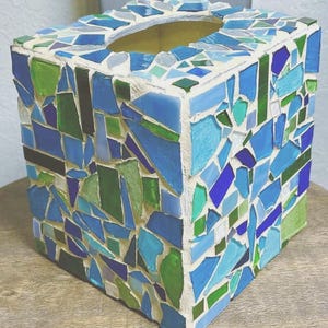 Op de afbeelding: Een vierkante tissueboxhouder versierd met een moza&iuml;ek van glasscherven in diverse tinten blauw, groen en wit. De stukjes zijn ingebed in een lichtgekleurde voegmortel, wat zorgt voor een getextureerd en veelkleurig oppervlak aan alle zijden.