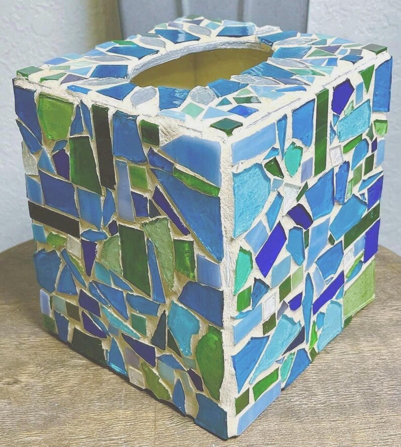Op de afbeelding: Een vierkante tissueboxhouder versierd met een moza&iuml;ek van glasscherven in diverse tinten blauw, groen en wit. De stukjes zijn ingebed in een lichtgekleurde voegmortel, wat zorgt voor een getextureerd en veelkleurig oppervlak aan alle zijden.