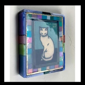 May include: A rectangular decorative box featuring a central image of a white cat on a dark blue background with the text A.J.D.G 1917. The frame is composed of small, multicolored glass tiles in shades of blue, pink, green, and purple with gold trim.