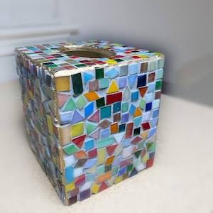 K&ouml;nnte beinhalten: Ein quadratischer Taschentuchbox-&Uuml;berzug, verziert mit einem bunten Mosaik aus kleinen Glasfliesen in verschiedenen Formen und Gr&ouml;&szlig;en. Die Kanten sind mit einer goldfarbenen Umrandung versehen und die Fliesen sind in wei&szlig;em Fugenm&ouml;rtel gesetzt.