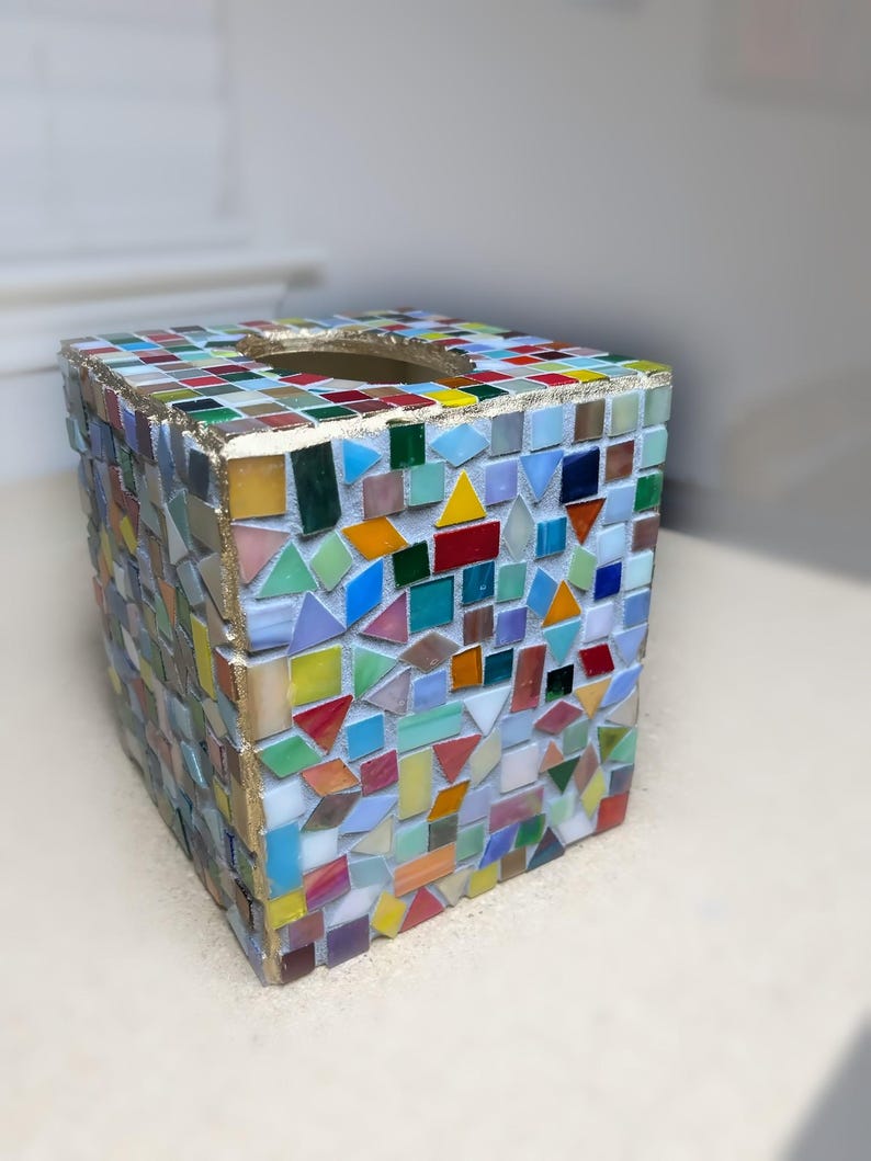 K&ouml;nnte beinhalten: Ein quadratischer Taschentuchbox-&Uuml;berzug, verziert mit einem bunten Mosaik aus kleinen Glasfliesen in verschiedenen Formen und Gr&ouml;&szlig;en. Die Kanten sind mit einer goldfarbenen Umrandung versehen und die Fliesen sind in wei&szlig;em Fugenm&ouml;rtel gesetzt.