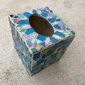 Op de afbeelding: Een vierkante tissueboxhouder versierd met een moza&iuml;ek van iriserende, blauwe en groene glasscherven in lichtgekleurde voegspecie. De bovenkant heeft een ovale opening en de zijkanten zijn bedekt met een dicht patroon van diverse glasstukjes.
