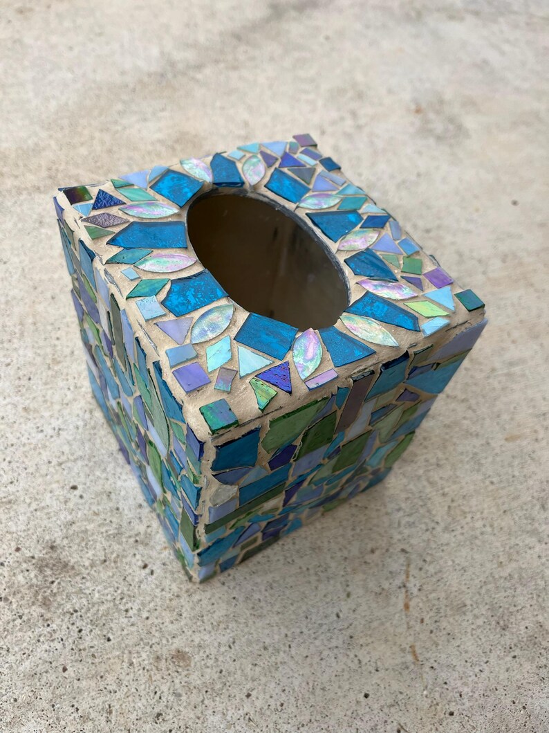 Op de afbeelding: Een vierkante tissueboxhouder versierd met een moza&iuml;ek van iriserende, blauwe en groene glasscherven in lichtgekleurde voegspecie. De bovenkant heeft een ovale opening en de zijkanten zijn bedekt met een dicht patroon van diverse glasstukjes.