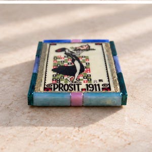 May include: A rectangular decorative tile featuring a central illustration of a figure in a black dress holding a tray, set against a checkered background. The text PROSIT 1911 is printed at the bottom. The tile is framed by blue and pink segments.