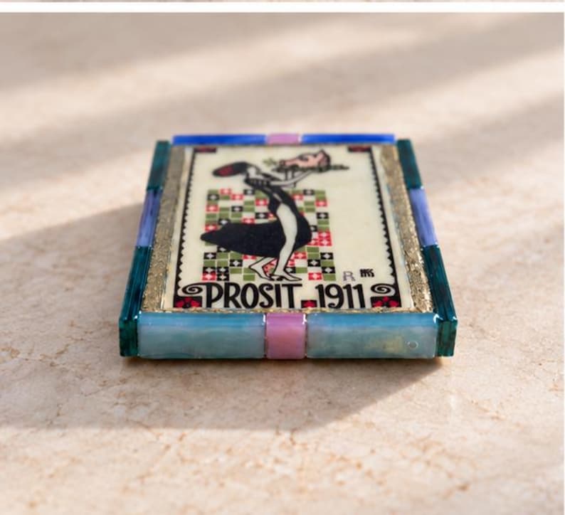 May include: A rectangular decorative tile featuring a central illustration of a figure in a black dress holding a tray, set against a checkered background. The text PROSIT 1911 is printed at the bottom. The tile is framed by blue and pink segments.