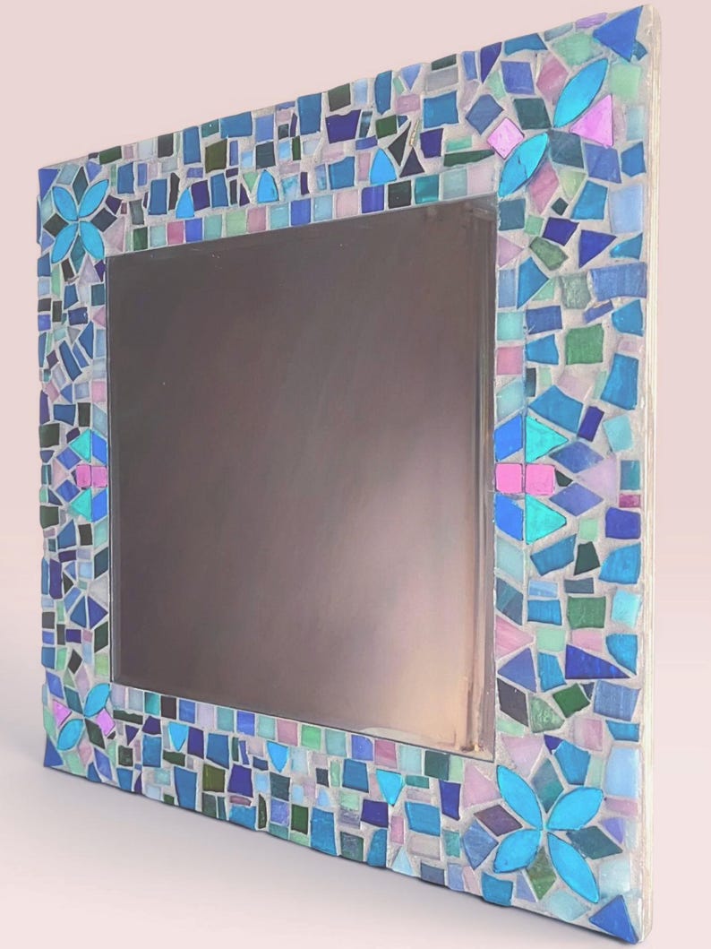 Puede incluir: Un espejo cuadrado con un marco de mosaico decorativo compuesto por peque&ntilde;as piezas de vidrio de formas irregulares en tonos azul, turquesa, verde y rosa claro. El marco incluye cuatro patrones florales distintos en las esquinas.