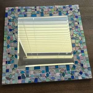 Op de afbeelding: Een vierkante wandspiegel met een decoratieve moza&iuml;eklijst bestaande uit talloze kleine, onregelmatig gevormde glazen tegels in diverse tinten blauw, lichtgroen, roze en iriserend wit, gerangschikt in een dicht, veelkleurig patroon.