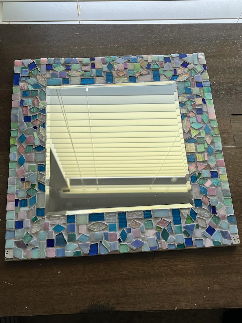 Op de afbeelding: Een vierkante wandspiegel met een decoratieve moza&iuml;eklijst bestaande uit talloze kleine, onregelmatig gevormde glazen tegels in diverse tinten blauw, lichtgroen, roze en iriserend wit, gerangschikt in een dicht, veelkleurig patroon.
