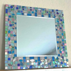 May include: A square wall mirror featuring a decorative frame composed of numerous small, multi-shaped glass tiles in shades of blue, turquoise, pink, and white, all set within a light gray grout that fills the spaces between the glass pieces.