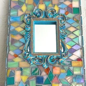 May include: A rectangular wall mirror featuring a central ornate turquoise and gold frame surrounded by a mosaic border of multicolored glass tiles in shades of yellow, blue, green, pink, and orange set in gray grout with a rectangular shape.
