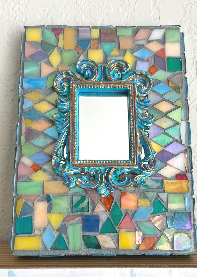 May include: A rectangular wall mirror featuring a central ornate turquoise and gold frame surrounded by a mosaic border of multicolored glass tiles in shades of yellow, blue, green, pink, and orange set in gray grout with a rectangular shape.