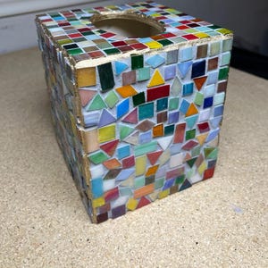 K&ouml;nnte beinhalten: Ein quadratischer Taschentuchbox-&Uuml;berzug, verziert mit einem bunten Mosaik aus kleinen Glasfliesen in verschiedenen geometrischen Formen. Die Kanten sind mit einem goldfarbenen Finish akzentuiert und die Oberseite hat eine runde &Ouml;ffnung.