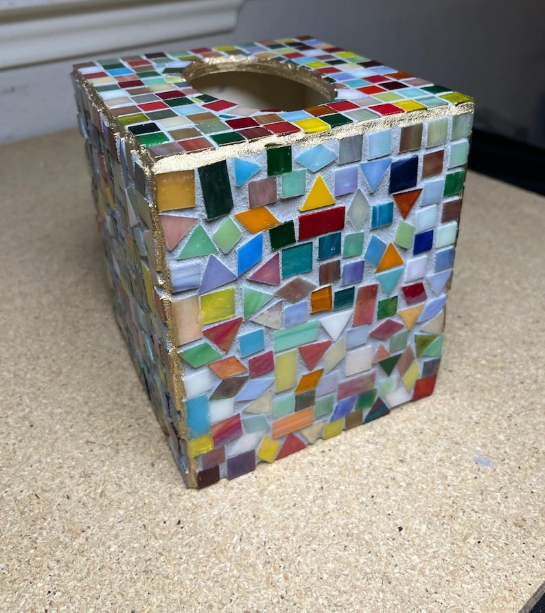 K&ouml;nnte beinhalten: Ein quadratischer Taschentuchbox-&Uuml;berzug, verziert mit einem bunten Mosaik aus kleinen Glasfliesen in verschiedenen geometrischen Formen. Die Kanten sind mit einem goldfarbenen Finish akzentuiert und die Oberseite hat eine runde &Ouml;ffnung.
