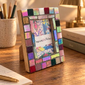 May include: A rectangular picture frame featuring a mosaic border made of small, multicolored, iridescent, and solid-colored square tiles. The frame holds a white insert with the text "pascaleRow" printed in a dark, stylized script font.