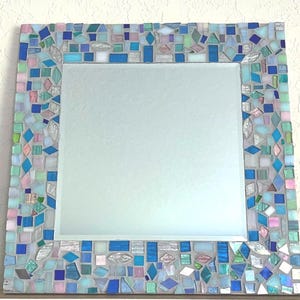 Op de afbeelding: Een vierkante wandspiegel met een decoratieve lijst bestaande uit talloze kleine, onregelmatig gevormde moza&iuml;ekglastegels in tinten blauw, lichtgroen, roze en wit, geplaatst in een lichtgrijze voegbasis rondom een centrale glazen spiegelplaat.