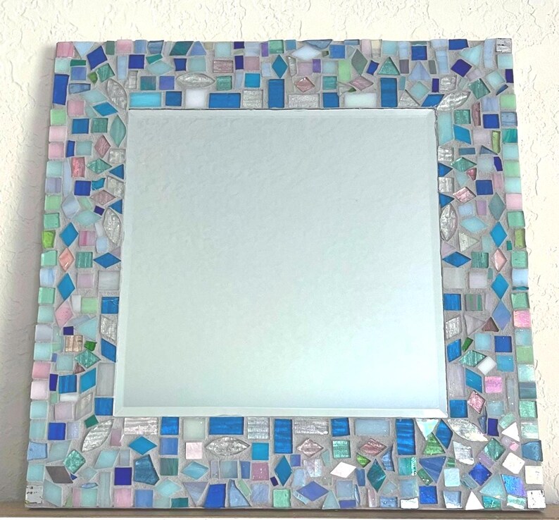 Op de afbeelding: Een vierkante wandspiegel met een decoratieve lijst bestaande uit talloze kleine, onregelmatig gevormde moza&iuml;ekglastegels in tinten blauw, lichtgroen, roze en wit, geplaatst in een lichtgrijze voegbasis rondom een centrale glazen spiegelplaat.