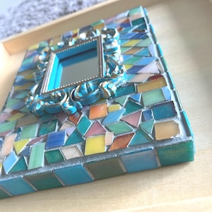 May include: A rectangular wall mirror featuring a central frame with ornate blue and silver detailing, surrounded by a mosaic of multi-colored glass tiles in shades of teal, blue, yellow, and pink, all set within a light-colored wooden frame.