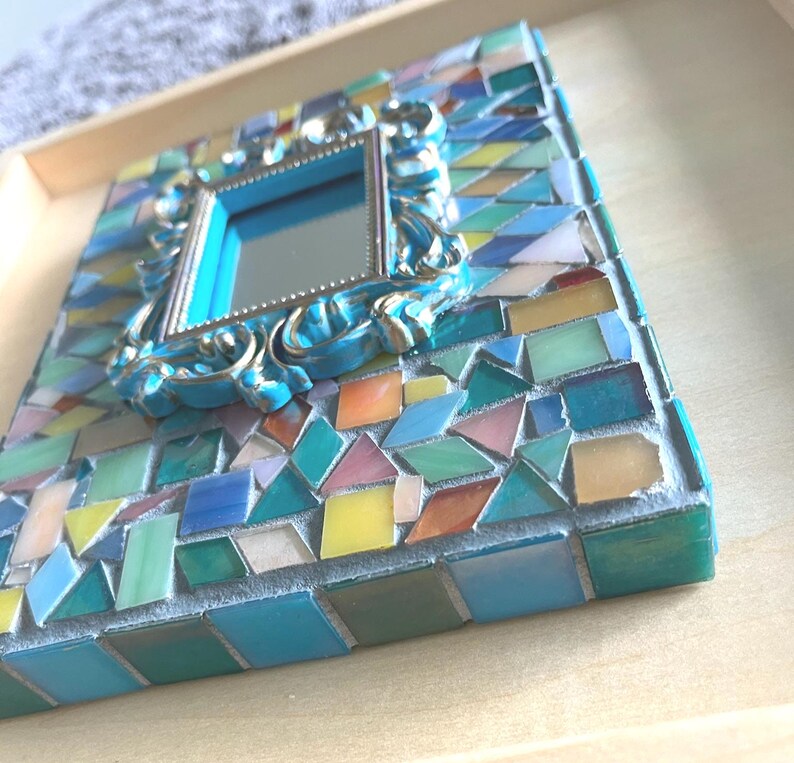 May include: A rectangular wall mirror featuring a central frame with ornate blue and silver detailing, surrounded by a mosaic of multi-colored glass tiles in shades of teal, blue, yellow, and pink, all set within a light-colored wooden frame.