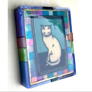 May include: A rectangular decorative box featuring a central image of a white cat on a teal and dark blue background with the text A.J.D.G 1917. The border is composed of small, multicolored glass tiles in shades of blue, pink, green, and purple.