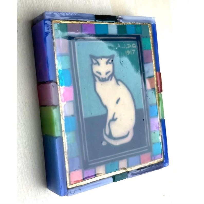 May include: A rectangular decorative box featuring a central image of a white cat on a teal and dark blue background with the text A.J.D.G 1917. The border is composed of small, multicolored glass tiles in shades of blue, pink, green, and purple.