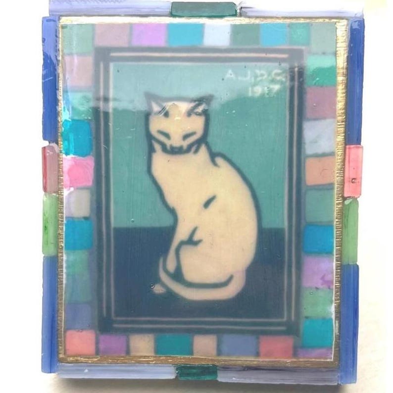 May include: A rectangular glass art piece featuring a central illustration of a cream-colored cat sitting against a teal and dark blue background. The image includes the text A.J.P.G. 1917. A border of small, multicolored glass tiles surrounds the center.