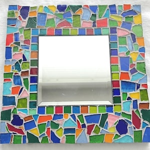 May include: A square wall mirror featuring a decorative frame composed of multi-colored glass mosaic tiles. The border includes a variety of irregular shapes and sizes in shades of blue, green, yellow, orange, red, and pink, set in white grout.