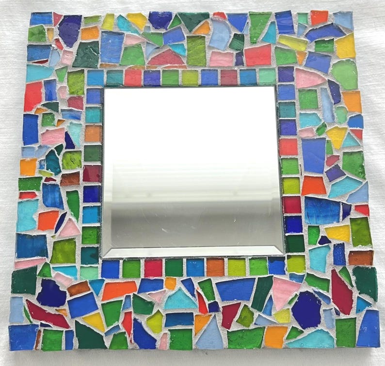 May include: A square wall mirror featuring a decorative frame composed of multi-colored glass mosaic tiles. The border includes a variety of irregular shapes and sizes in shades of blue, green, yellow, orange, red, and pink, set in white grout.