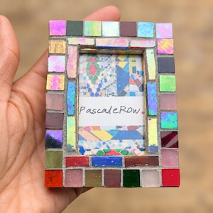 May include: A rectangular picture frame decorated with a mosaic of small, multi-colored glass tiles in shades of red, blue, green, yellow, and iridescent tones. The center features a white insert with the text PascaleRow printed in black ink.