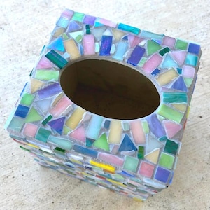 May include: A rectangular tissue box cover featuring a mosaic design made of small, multi-colored glass tiles in shades of pink, blue, green, yellow, and purple. The tiles are arranged in a random pattern and set in light gray grout.