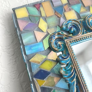 May include: A rectangular mirror frame featuring a mosaic border of iridescent, multi-colored glass tiles in shades of yellow, blue, pink, and green. The inner edge is defined by an ornate, scrolled blue and gold resin border with bead detailing.