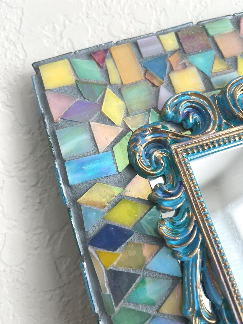 May include: A rectangular mirror frame featuring a mosaic border of iridescent, multi-colored glass tiles in shades of yellow, blue, pink, and green. The inner edge is defined by an ornate, scrolled blue and gold resin border with bead detailing.