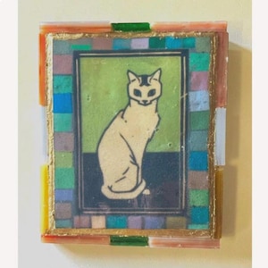 May include: A rectangular wall art piece featuring a central illustration of a seated cream-colored cat against a green and dark blue background. The artwork is framed by a border of small, multicolored glass tiles in shades of blue, green, and orange.
