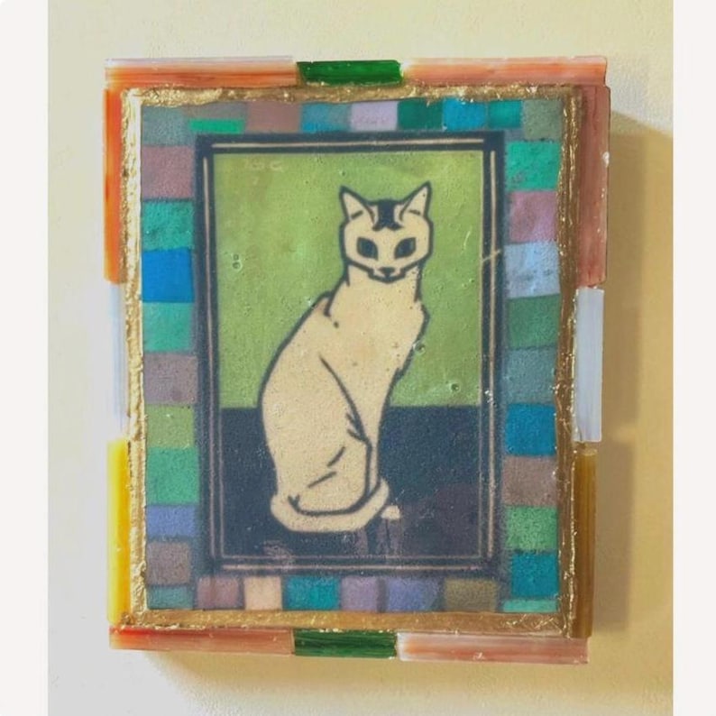 May include: A rectangular wall art piece featuring a central illustration of a seated cream-colored cat against a green and dark blue background. The artwork is framed by a border of small, multicolored glass tiles in shades of blue, green, and orange.