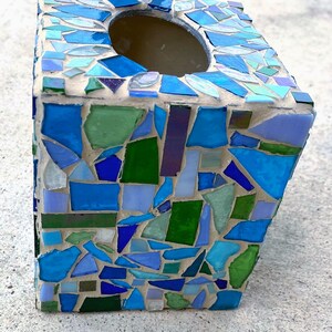 Op de afbeelding: Een vierkante tissueboxhouder met een moza&iuml;ekontwerp van diverse glasscherven in tinten blauw, groen en lichtpaars. De glasstukjes zijn ingebed in lichtgekleurde voegspecie, wat zorgt voor een textuurrijk oppervlak met een ronde opening aan de bovenkant.