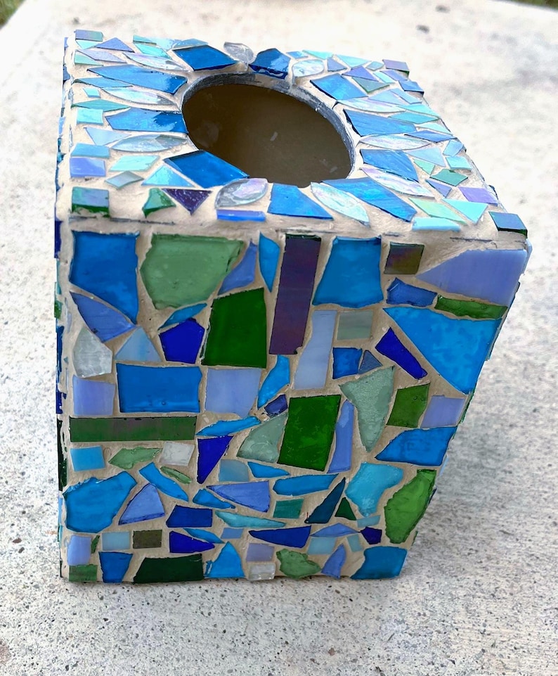 Op de afbeelding: Een vierkante tissueboxhouder met een moza&iuml;ekontwerp van diverse glasscherven in tinten blauw, groen en lichtpaars. De glasstukjes zijn ingebed in lichtgekleurde voegspecie, wat zorgt voor een textuurrijk oppervlak met een ronde opening aan de bovenkant.