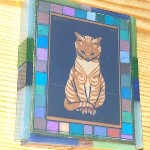 Pode incluir: Uma moldura de vidro retangular com uma ilustra&ccedil;&atilde;o central de um gato malhado laranja sentado sobre um fundo azul escuro. A borda &eacute; composta por pequenos azulejos de vidro multicoloridos em tons de azul, verde e roxo. O texto AJDC 1818 &eacute; vis&iacute;vel.