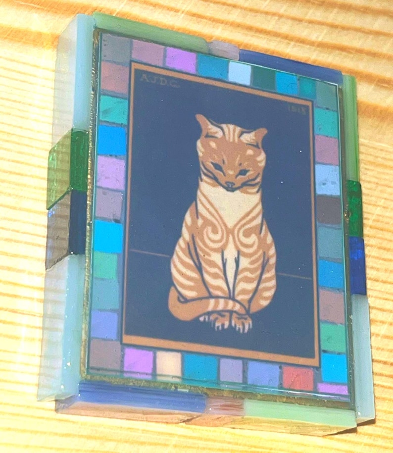 Pode incluir: Uma moldura de vidro retangular com uma ilustra&ccedil;&atilde;o central de um gato malhado laranja sentado sobre um fundo azul escuro. A borda &eacute; composta por pequenos azulejos de vidro multicoloridos em tons de azul, verde e roxo. O texto AJDC 1818 &eacute; vis&iacute;vel.