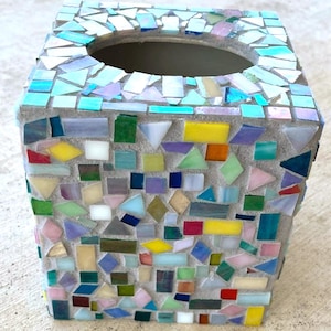 May include: A square tissue box cover featuring a mosaic design made of various small, multi-colored glass pieces set in light gray grout. The surface includes shades of blue, green, yellow, red, and white, with an oval opening at the top.