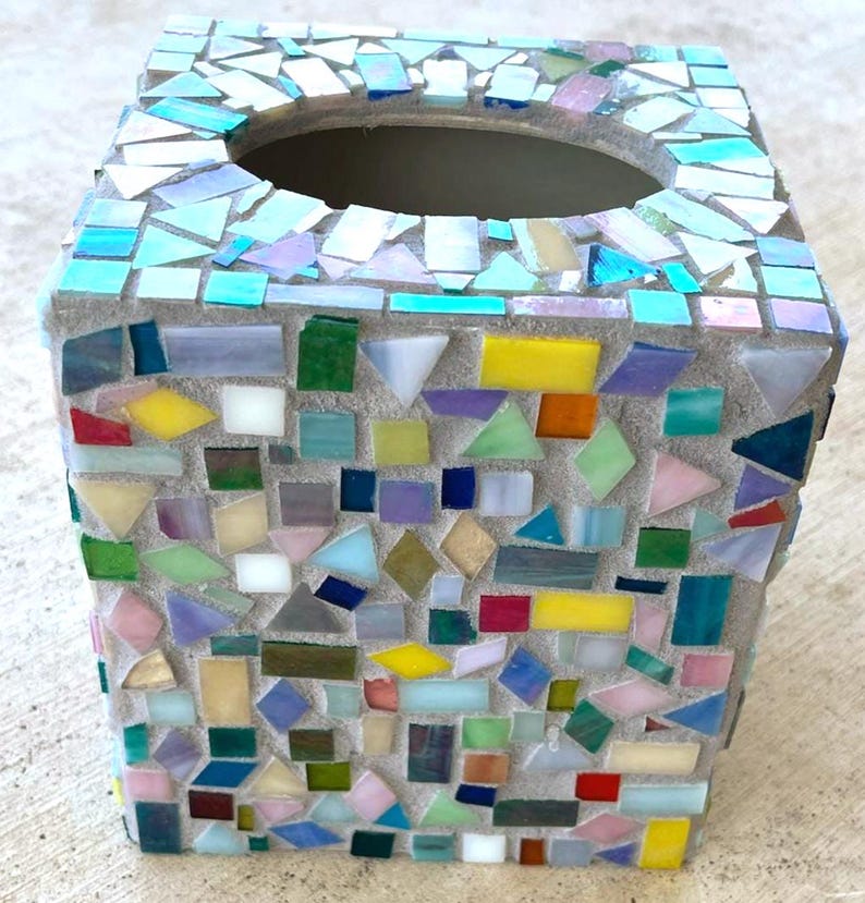May include: A square tissue box cover featuring a mosaic design made of various small, multi-colored glass pieces set in light gray grout. The surface includes shades of blue, green, yellow, red, and white, with an oval opening at the top.