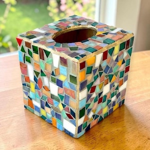 May include: A square tissue box cover decorated with a multi-coloured mosaic pattern of small glass tiles in shades of red, blue, green, yellow, and white. The top features an oval opening with a gold-toned border, and the tiles are set in grey grout.