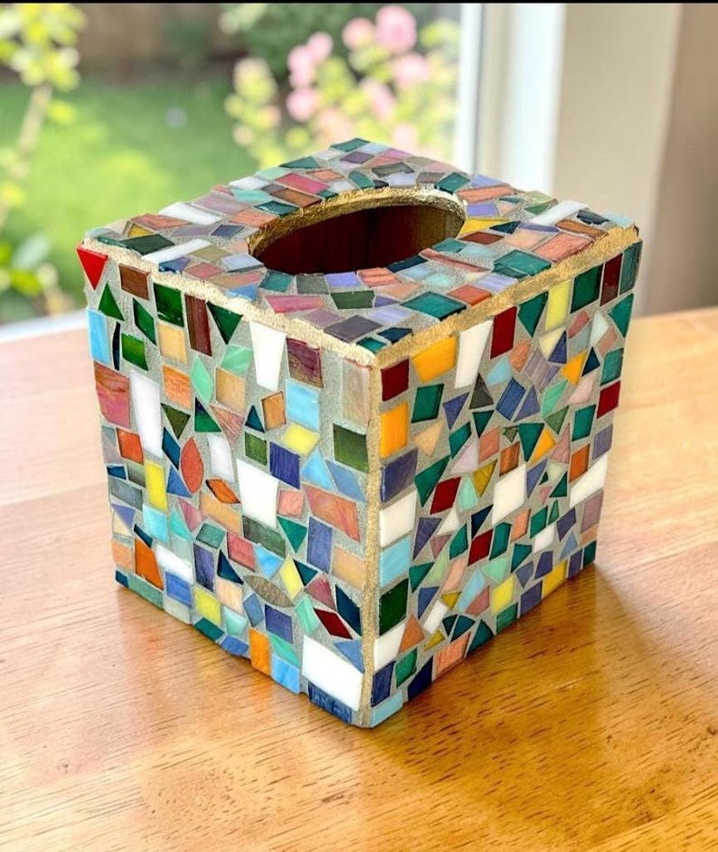 May include: A square tissue box cover decorated with a multi-coloured mosaic pattern of small glass tiles in shades of red, blue, green, yellow, and white. The top features an oval opening with a gold-toned border, and the tiles are set in grey grout.