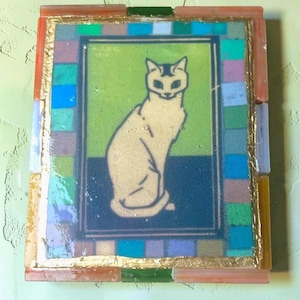 May include: A rectangular wall art piece featuring a central illustration of a seated cat in cream against a green and dark blue background. The artwork is framed by a border of multicolored glass tiles and a thin gold-colored metallic trim.