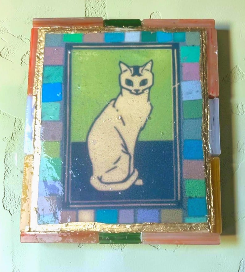 May include: A rectangular wall art piece featuring a central illustration of a seated cat in cream against a green and dark blue background. The artwork is framed by a border of multicolored glass tiles and a thin gold-colored metallic trim.