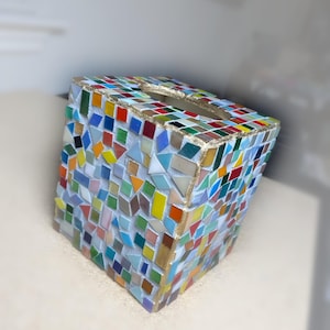 K&ouml;nnte beinhalten: Ein quadratischer Taschentuchbox-Bezug, verziert mit einem bunten Mosaikmuster aus kleinen, unregelm&auml;&szlig;igen Glasfliesen in Rot-, Blau-, Gr&uuml;n-, Gelb- und Oranget&ouml;nen. Die Kanten sind mit einer goldfarbenen Verzierung versehen und oben befindet sich eine ovale &Ouml;ffnung.