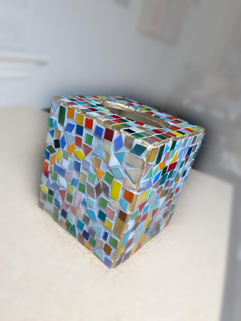 K&ouml;nnte beinhalten: Ein quadratischer Taschentuchbox-Bezug, verziert mit einem bunten Mosaikmuster aus kleinen, unregelm&auml;&szlig;igen Glasfliesen in Rot-, Blau-, Gr&uuml;n-, Gelb- und Oranget&ouml;nen. Die Kanten sind mit einer goldfarbenen Verzierung versehen und oben befindet sich eine ovale &Ouml;ffnung.