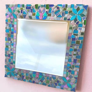 Puede incluir: Un espejo cuadrado con un marco de mosaico decorativo compuesto por peque&ntilde;as piezas de vidrio de formas irregulares en tonos turquesa, azul cobalto, verde claro y lavanda suave, con cuatro acentos florales en las esquinas.