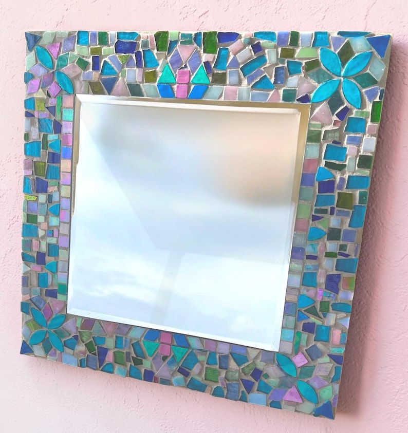 Puede incluir: Un espejo cuadrado con un marco de mosaico decorativo compuesto por peque&ntilde;as piezas de vidrio de formas irregulares en tonos turquesa, azul cobalto, verde claro y lavanda suave, con cuatro acentos florales en las esquinas.