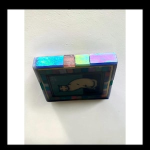 May include: A square decorative tile featuring a central illustration of a cream-colored cat against a dark background. The tile is framed by a border of small, iridescent, multi-colored glass squares in shades of blue, green, and purple.