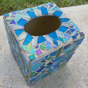 Op de afbeelding: Een vierkante tissueboxhouder versierd met een moza&iuml;ek van diverse blauwe, groene en iriserende glasstukjes in lichtbeige voegmiddel. De bovenkant heeft een ovale opening en de zijkanten zijn bedekt met geometrische scherven van gekleurd glas.