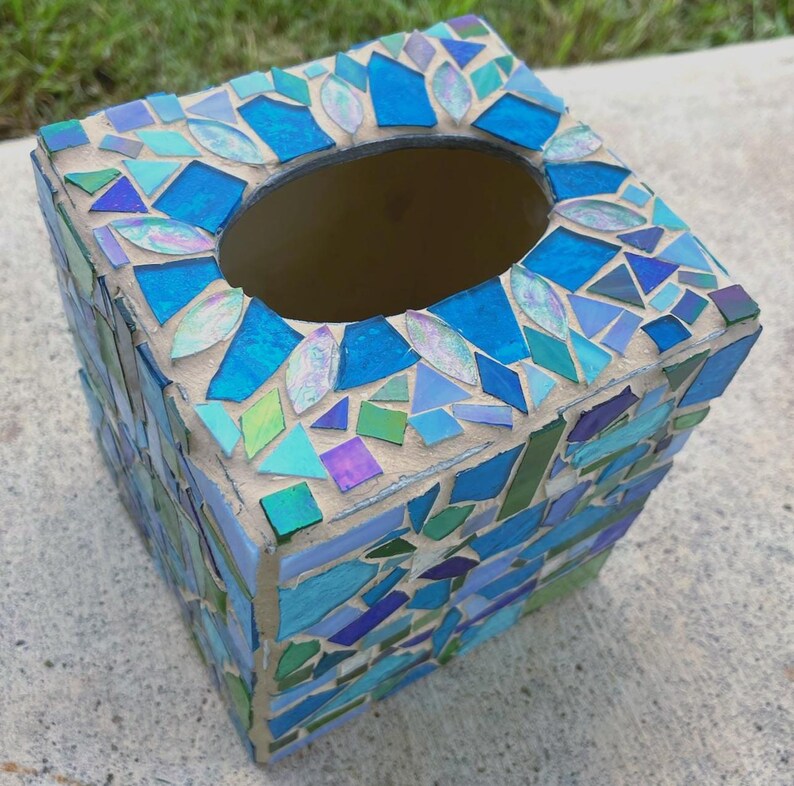 Op de afbeelding: Een vierkante tissueboxhouder versierd met een moza&iuml;ek van diverse blauwe, groene en iriserende glasstukjes in lichtbeige voegmiddel. De bovenkant heeft een ovale opening en de zijkanten zijn bedekt met geometrische scherven van gekleurd glas.